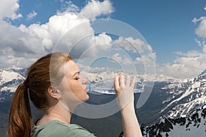 Young woman drink water from plastic bottle in the mountains on the snow peaks background