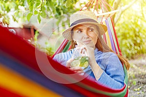 Young woman drinking cocktail while lying in comfortable hammock at green garden