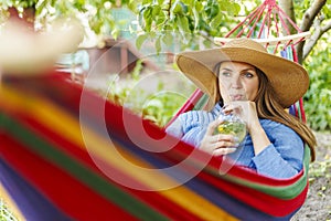 Young woman drinking cocktail while lying in comfortable hammock at green garden