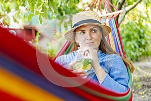 Young woman drinking cocktail while lying in comfortable hammock at green garden