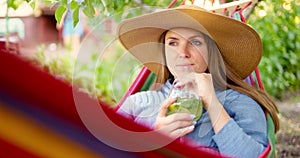 Young woman drinking cocktail while lying in comfortable hammock at green garden