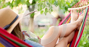 Young woman drinking cocktail while lying in comfortable hammock at green garden