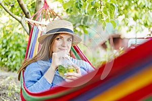 Young woman drinking cocktail while lying in comfortable hammock at green garden