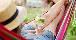 Young woman drinking cocktail while lying in comfortable hammock at green garden