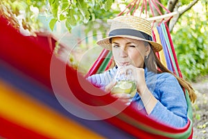 Young woman drinking cocktail while lying in comfortable hammock at green garden