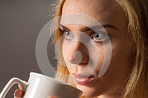 Young woman drinking coffee. Blonde girl with white tea cup on a gray background.