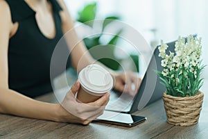 Young woman drinking coffee from disposable cup and using computer laptop on work desk