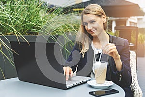Young woman drinking coffee latte at cafe outdoor terrace and working with laptop. freelance remote work