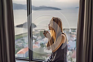 Young woman is drinking coffee in the morning on the background of a window with a sea view