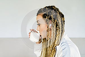 A young woman drinking coffee is in profile with braids in all her hair wearing a very large golden ring and headphones