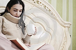 Young woman drinking coffee and reading a book in the bed