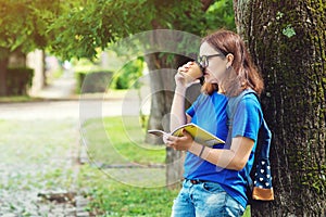 Young woman drinking coffee and reading a book at summer park. Student girl enjoys outdoor recreation. College girl holding coffee