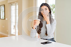 Young woman drinking a cup of coffee at home cover mouth with hand shocked with shame for mistake, expression of fear, scared in