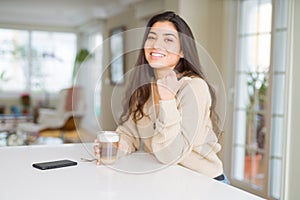 Young woman drinking a cup of coffee at home pointing and showing with thumb up to the side with happy face smiling