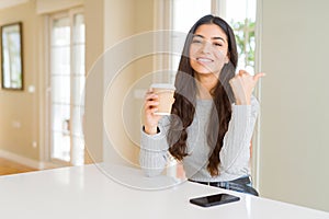 Young woman drinking a cup of coffee at home pointing and showing with thumb up to the side with happy face smiling