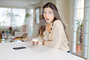 Young woman drinking a cup of coffee at home scared in shock with a surprise face, afraid and excited with fear expression