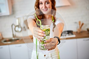 Young woman drinking fresh water with cucumber, lemon and leaves of mint from glass in the kitchen. Healthy Lifestyle and Eating.