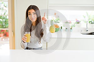 Young woman drinking a glass of fresh orange juice surprised with an idea or question pointing finger with happy face, number one