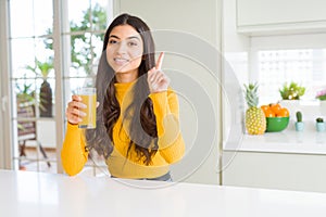 Young woman drinking a glass of fresh orange juice surprised with an idea or question pointing finger with happy face, number one