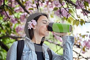 Young woman drinking from green bottle on the background of blooming sakura