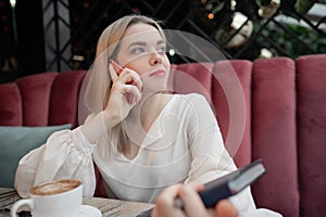 Young woman drinking hot coffee and talking on phone in cafe holding notepad