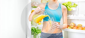 Young woman drinking juice and staing near the fridge full of health food.