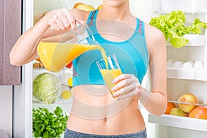 Young woman drinking juice and staing near the fridge full of health food.