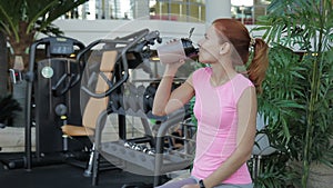 Young woman is drinking protein cocktail while sitting in gym.