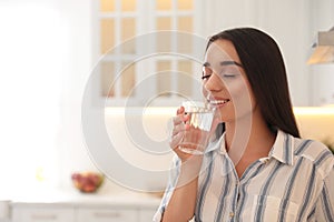Young woman drinking pure water from glass in kitchen. Space for text