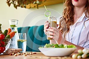Young woman drinking smoothie in the beautiful interior with green flowers on the background and fresh fruits and vegetables on