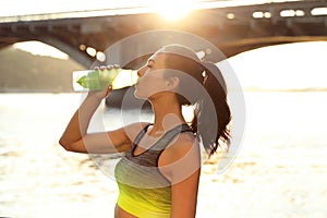 Young woman drinking water after running near river on sunny day