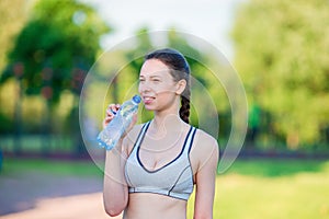 Young woman drinking water after running outside. Female fitness model training outside in the park. Concept of healthy