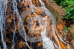 A young woman drinks clean water from a bottle in the jungle with a waterfall in the background.