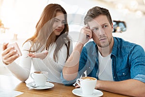 Young woman drinks coffee with her husband, she shows him photoes on the phone while he turned around looking tired, digital