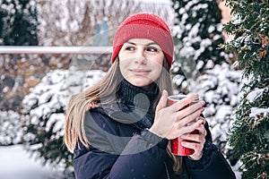 A young woman drinks a hot drink from a red thermal cup in winter.