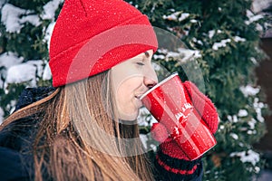 A young woman drinks a hot drink from a red thermal cup in winter.