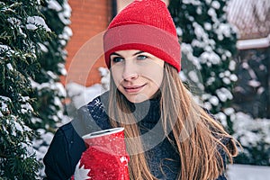 A young woman drinks a hot drink from a red thermal cup in winter.