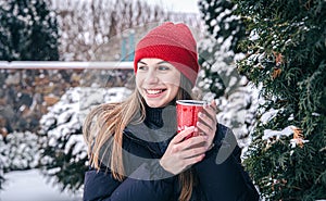 A young woman drinks a hot drink from a red thermal cup in winter.