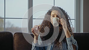 Young woman drinks tea with a cookie sitting on a couch in the sunny morning