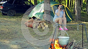 A young woman drinks tea near a tent and watches a bowler hanging over a fire.