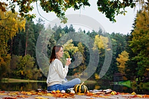 A young woman drinks tea from a white mug on a wooden bridge on the lake. Rest in the warm autumn in nature