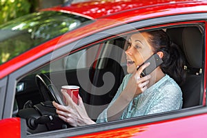 A young woman driver in a car with a cup of coffee in her hands speaks on a phone.