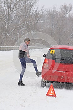 Young woman driver checks tire of  car on road in  blizzard and bad weather. Triangular accident warning sign stands on the