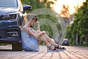 Young woman driver sitting beside her broken car waiting for help. Vehicle problems concept