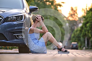 Young woman driver sitting beside her broken car waiting for help. Vehicle problems concept