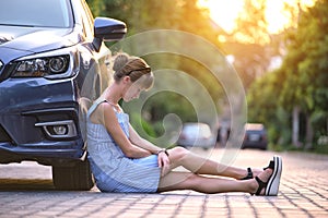 Young woman driver sitting beside her broken car waiting for help. Vehicle problems concept