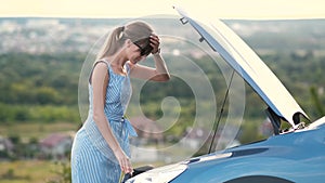 Young woman driver standing near a broken car with open hood having trouble with her vehicle