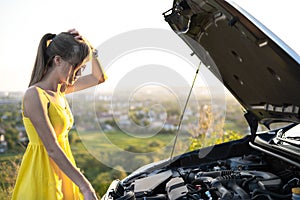 Young woman driver standing near her car with open hood having motor problem