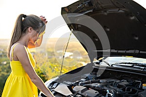 Young woman driver standing near her car with popped hood having engine problems