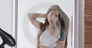 Young woman drying her hair with dryer, looking at mirror